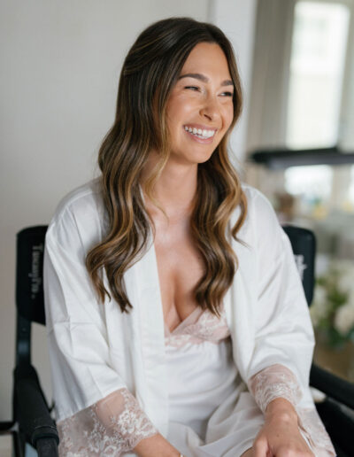 A woman with long brown wavy hair, wearing a white robe with lace cuffs, sits and smiles indoors.