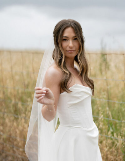 A woman in a white strapless wedding dress and veil stands outdoors near a wire fence, with tall grass and a cloudy sky in the background.