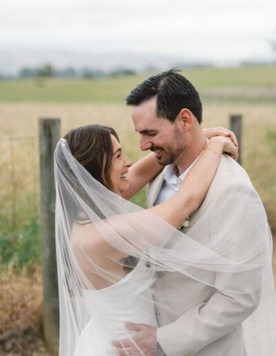 A bride and groom embrace outdoors, smiling at each other, with the bride’s veil draped over both of them and a field in the background.