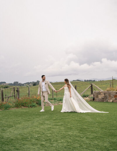 A bride and groom hold hands and walk on a grassy field. The bride wears a white dress with a long veil, and the groom wears a light-colored suit. A stone wall and greenery are in the background.