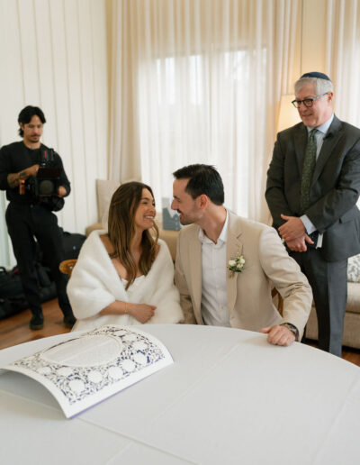 A bride and groom sit smiling at a table with a decorative document, as an officiant and a photographer stand nearby in a bright room.