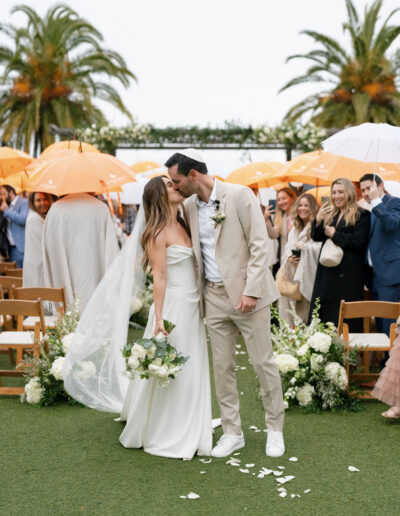 A bride and groom kiss at an outdoor wedding ceremony, surrounded by guests holding orange and white umbrellas, with palm trees in the background.