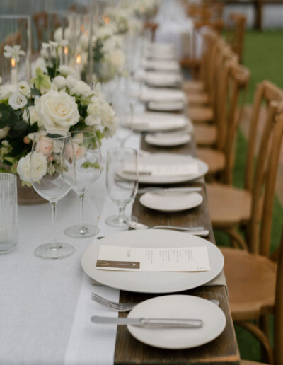A long table set for an event with white plates, menus, glassware, silverware, and floral centerpieces featuring white roses on a white tablecloth.