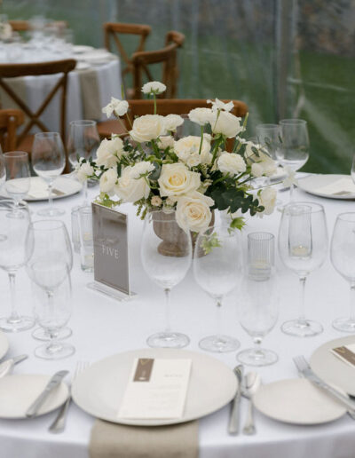 A round table set for an event with white tablecloth, glassware, plates, menus, beige napkins, and a centerpiece of white roses and greenery. Wooden chairs surround the table.