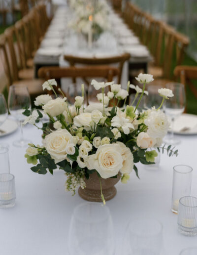 A white floral centerpiece with roses and greenery sits on a long, formally set dining table with wooden chairs and glassware.