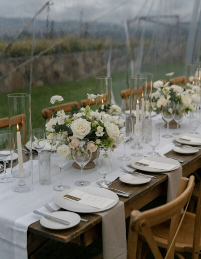 A long wooden table set for a formal event with white flowers, glass candle holders, menus, plates, and glasses on a white table runner, under a transparent tent.