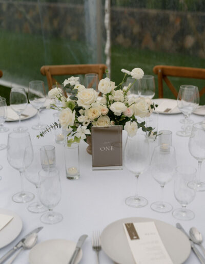 A round table set for an event with white flowers in a vase, a table number card, clear glassware, white plates, cutlery, and menus on a white tablecloth.
