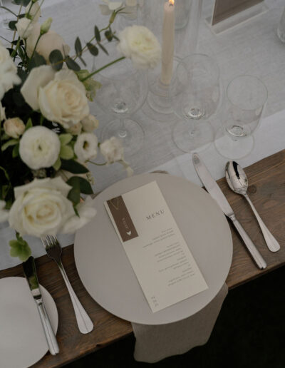 A neutral-toned table setting with white flowers, candles, glassware, cutlery, and a printed menu card placed on a round plate.