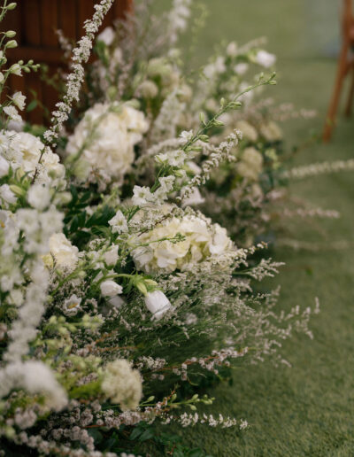 White flowers and greenery arranged closely together sit on a patch of green grass, with wooden furniture partially visible in the background.