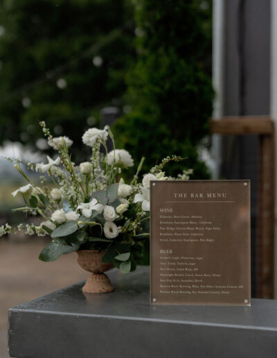 A floral arrangement in a brown vase sits next to a clear sign displaying a bar menu with wine and beer options on a metal countertop.