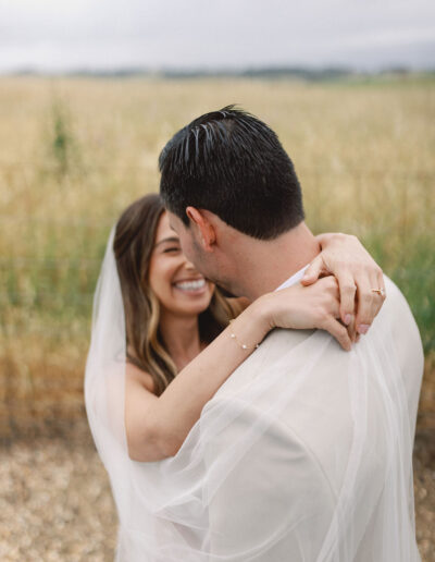 A bride and groom embrace outdoors, smiling and wearing wedding attire, with a field and overcast sky in the background.