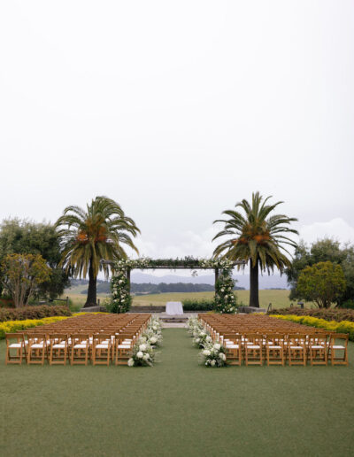 Outdoor wedding ceremony setup with rows of wooden chairs, white floral arrangements lining the aisle, and two palm trees framing an arbor on a lawn.