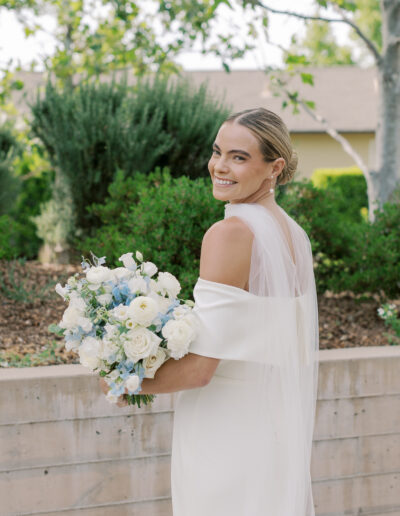 A bride in an off-shoulder white dress holds a bouquet of white and light blue flowers, smiling over her shoulder in the lush gardens of a Solage Calistoga wedding.