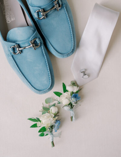 Light blue loafers, a white necktie with cufflinks, and two small boutonnieres with white and blue flowers arranged on a white surface—perfect details for a Solage Calistoga wedding.