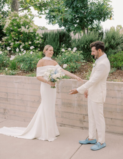 A bride in an off-shoulder white gown holds a bouquet and smiles at the groom in a white suit and blue shoes, standing outdoors near greenery at their Solage Calistoga wedding.