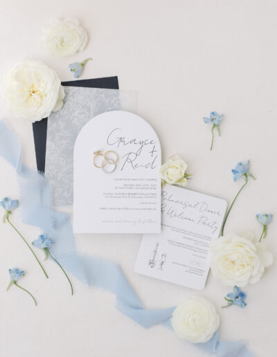 Elegant Solage Calistoga wedding invitation suite featuring white and translucent cards, gold rings, blue ribbon, and white and blue flowers beautifully arranged on a light surface.