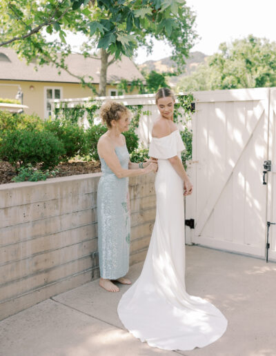 A woman in a light blue dress helps a bride in an off-shoulder white gown adjust her dress outdoors by a white fence on a sunny Solage Calistoga wedding day.
