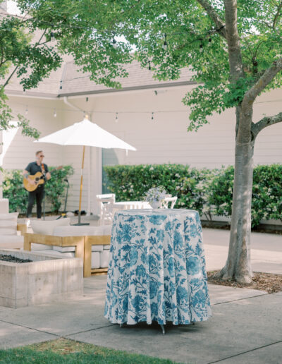 A round table with a blue floral tablecloth stands on a patio; nearby, a man plays guitar under a white umbrella, evoking the relaxed elegance of a Solage Calistoga wedding, surrounded by trees and outdoor seating.