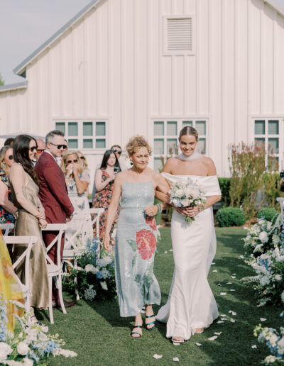 A bride in a white dress walks down an outdoor aisle with an older woman in a floral dress, surrounded by guests at a stunning Solage Calistoga wedding in front of a beautiful white building.