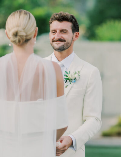 A man in a white suit with a boutonniere smiles at a woman in a white dress and veil during an outdoor Solage Calistoga wedding ceremony.