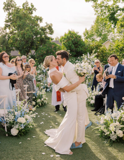 A newlywed couple kisses while walking down an outdoor aisle lined with flowers at their Solage Calistoga wedding, surrounded by applauding guests capturing the moment.