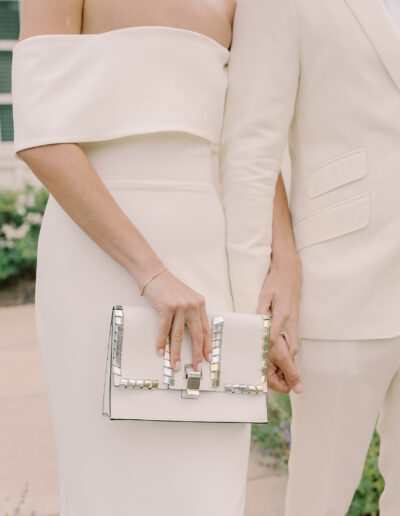 Two people dressed in elegant, off-white formal attire stand side by side at a Solage Calistoga wedding, one holding a white clutch with metallic accents.