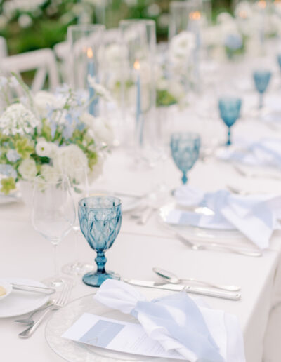 Elegant outdoor table setting at a Solage Calistoga wedding with white linens, blue glass goblets, white floral arrangements, candles in glass holders, and neatly folded napkins on clear plates.