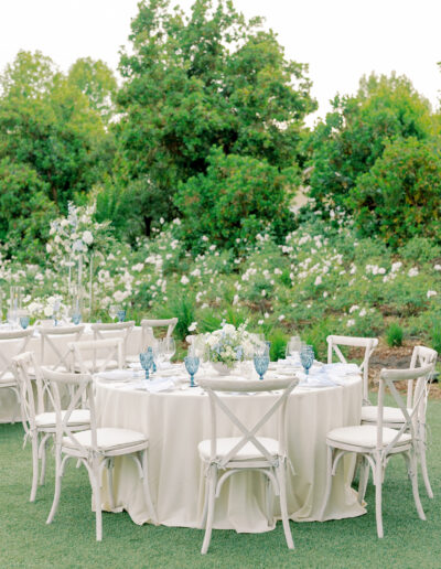 Round table covered with a white tablecloth, set with blue glassware and white floral centerpieces, surrounded by white chairs in an outdoor garden setting perfect for a Solage Calistoga wedding.