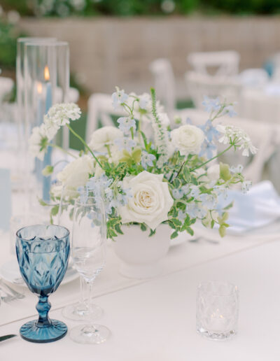 Elegant table setting at a Solage Calistoga wedding with a white floral centerpiece, blue glass goblet, clear wine and water glasses, a votive candle, and a tall glass candle holder atop a crisp white tablecloth.