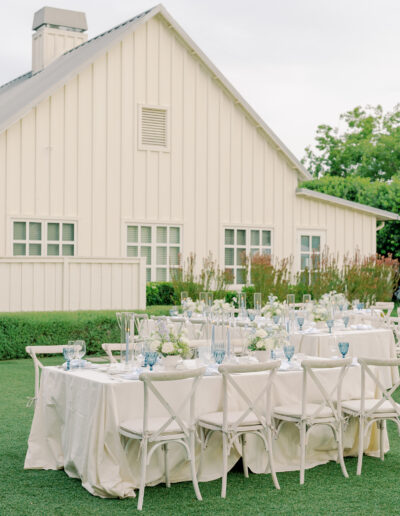 Two long tables with white tablecloths and blue glassware are set up outdoors on grass in front of a cream-colored building, capturing the elegant charm of a Solage Calistoga wedding.