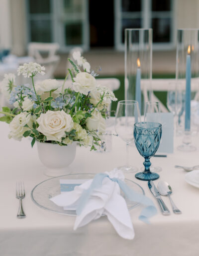 Table setting at a Solage Calistoga wedding with white flowers in a vase, blue glass goblet, clear plate, blue and white napkin, candles, and silverware on a white tablecloth.