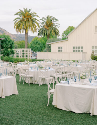 Outdoor Solage Calistoga wedding reception setup with long tables covered in white linens, blue glassware, and floral arrangements, set on grass near a white barn and palm trees.