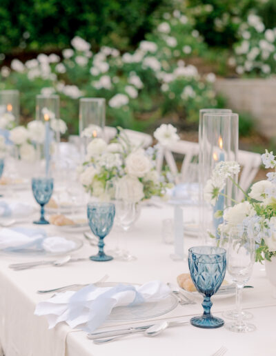 An elegant Solage Calistoga wedding outdoor table setting featuring white linens, blue glassware, white floral centerpieces, lit candles in tall holders, and neatly arranged cutlery and napkins.