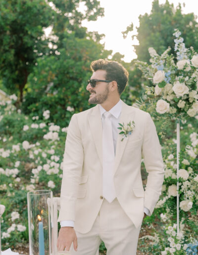 A man in a cream suit and sunglasses stands outdoors near a floral arrangement, surrounded by white roses and greenery at a Solage Calistoga wedding.