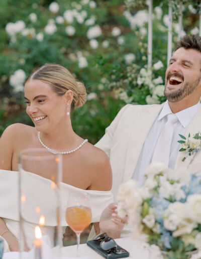 A bride and groom sit together at a decorated table outdoors at their Solage Calistoga wedding, smiling and laughing, surrounded by flowers and candles.
