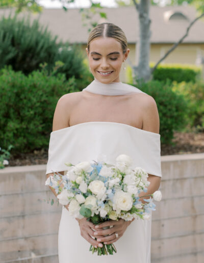 A woman in an off-shoulder white dress holds a bouquet of white and light blue flowers, standing outdoors by lush greenery and a low stone wall at a Solage Calistoga wedding.