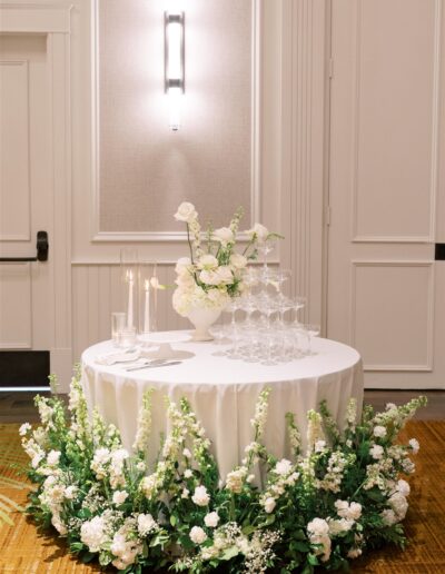 Round table with white tablecloth, surrounded by white and green floral arrangements, holds stacked champagne glasses, candles, and a white flower centerpiece in an elegant indoor setting.