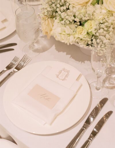 A formal place setting with a white plate, folded napkin, name card, silver cutlery, water glass, champagne flute, and a floral centerpiece with white and cream flowers.