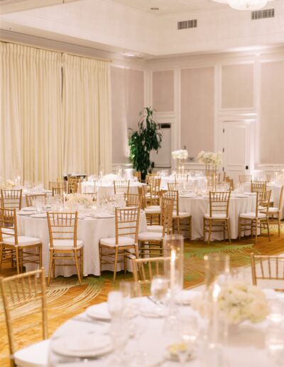 A banquet hall with round tables set for an event, featuring white tablecloths, gold chairs, floral centerpieces, and soft lighting.