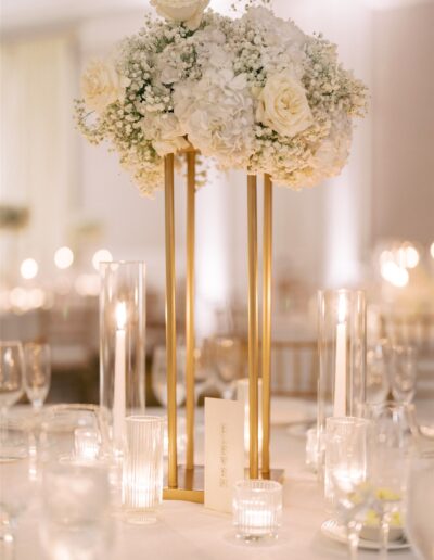 Tall gold centerpiece with white roses, hydrangeas, and baby's breath, surrounded by glass candles and table number on an elegant, softly lit table.