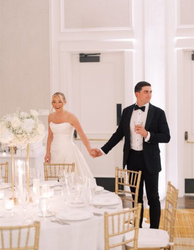 A bride and groom, dressed in formal wedding attire, hold hands while entering a decorated reception hall with round tables and gold chairs.