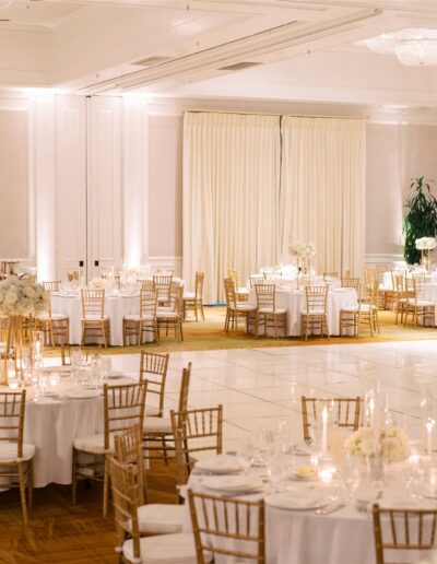 Elegant banquet hall with round tables set for an event, featuring white linens, gold chairs, floral centerpieces, and a large empty dance floor.