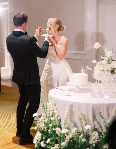 A bride and groom stand by a table with a wedding cake and flowers, feeding each other cake during their reception.