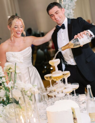 A woman in a white dress and a man in a tuxedo pour champagne into a tower of glasses at a decorated table with white flowers and a cake.