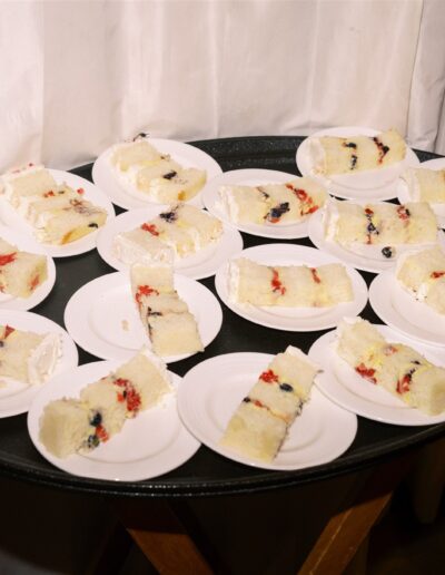 A round tray holds several small white plates, each with a slice of layered white cake filled with berries and cream. The tray is set against a white curtain background.