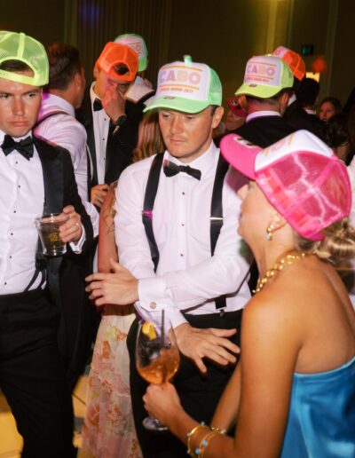 Several people dressed in formal attire and colorful neon hats dance and socialize at a lively indoor event or party.