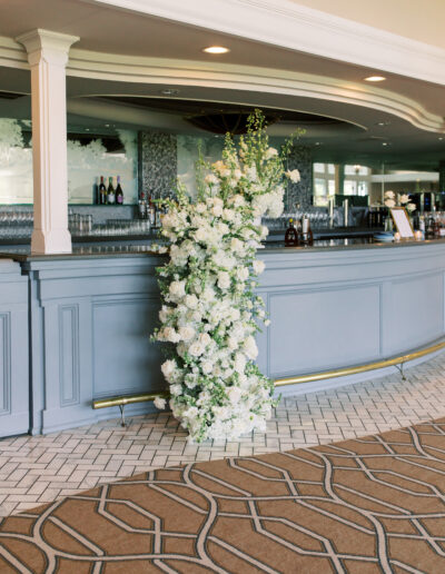 A tall vertical arrangement of white flowers with greenery stands in front of a gray bar counter in a spacious, well-lit room with patterned carpet and tiled floor.