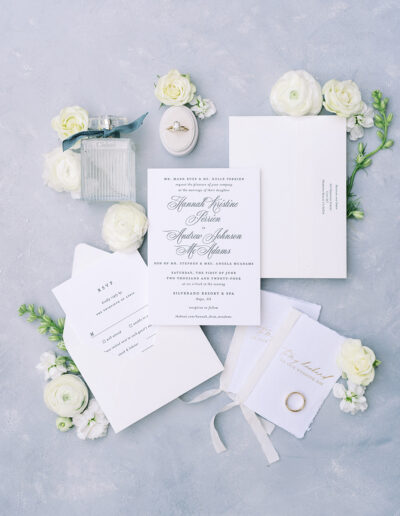 Flat lay of elegant wedding invitations, RSVP card, two vow books, a ring box with an engagement ring, a perfume bottle, and white flowers on a light blue textured surface.