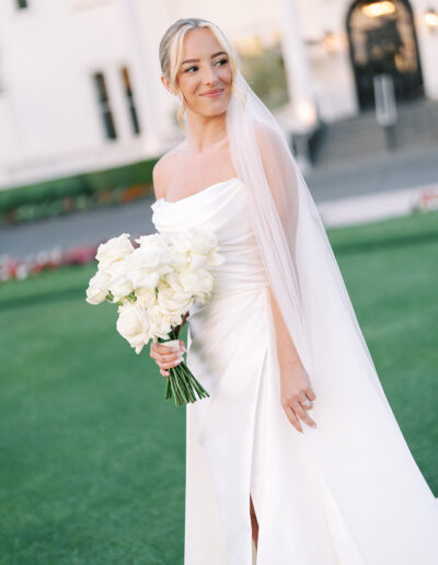 A bride in a white dress and veil stands outdoors holding a bouquet of white roses, with a building in the background.