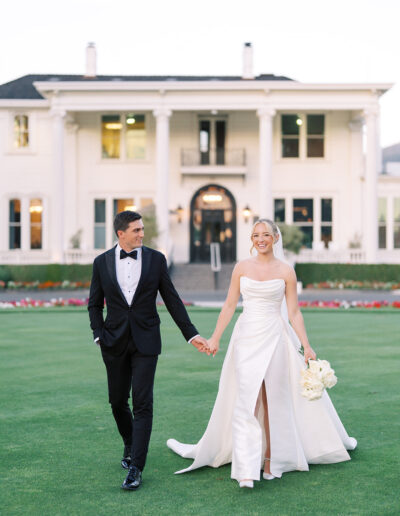 A bride and groom in formal attire walk hand in hand on a lawn in front of a large white house.